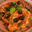 Petti di pollo alla mediterranea con sugo di pomodoro, olive nere fresche, capperi e foglie di basilico serviti su un piatto di terracotta.