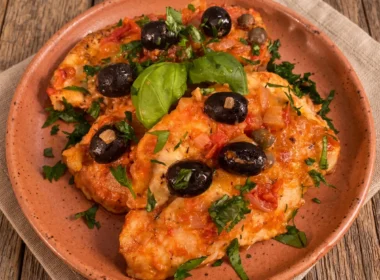 Petti di pollo alla mediterranea con sugo di pomodoro, olive nere fresche, capperi e foglie di basilico serviti su un piatto di terracotta.