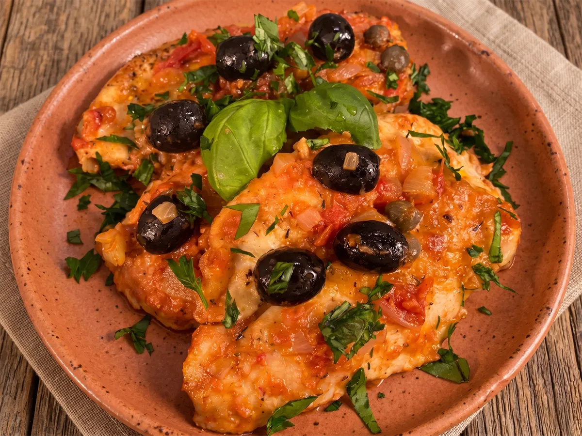 Petti di pollo alla mediterranea con sugo di pomodoro, olive nere fresche, capperi e foglie di basilico serviti su un piatto di terracotta.