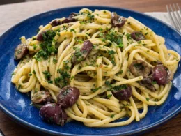 Un piatto in ceramica blu con spaghetti alla Cicerenella conditi con olive nere, burro, limone e prezzemolo fresco tritato, appoggiato su un tavolo di legno