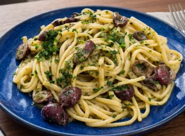 Un piatto in ceramica blu con spaghetti alla Cicerenella conditi con olive nere, burro, limone e prezzemolo fresco tritato, appoggiato su un tavolo di legno