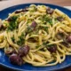 Un piatto in ceramica blu con spaghetti alla Cicerenella conditi con olive nere, burro, limone e prezzemolo fresco tritato, appoggiato su un tavolo di legno